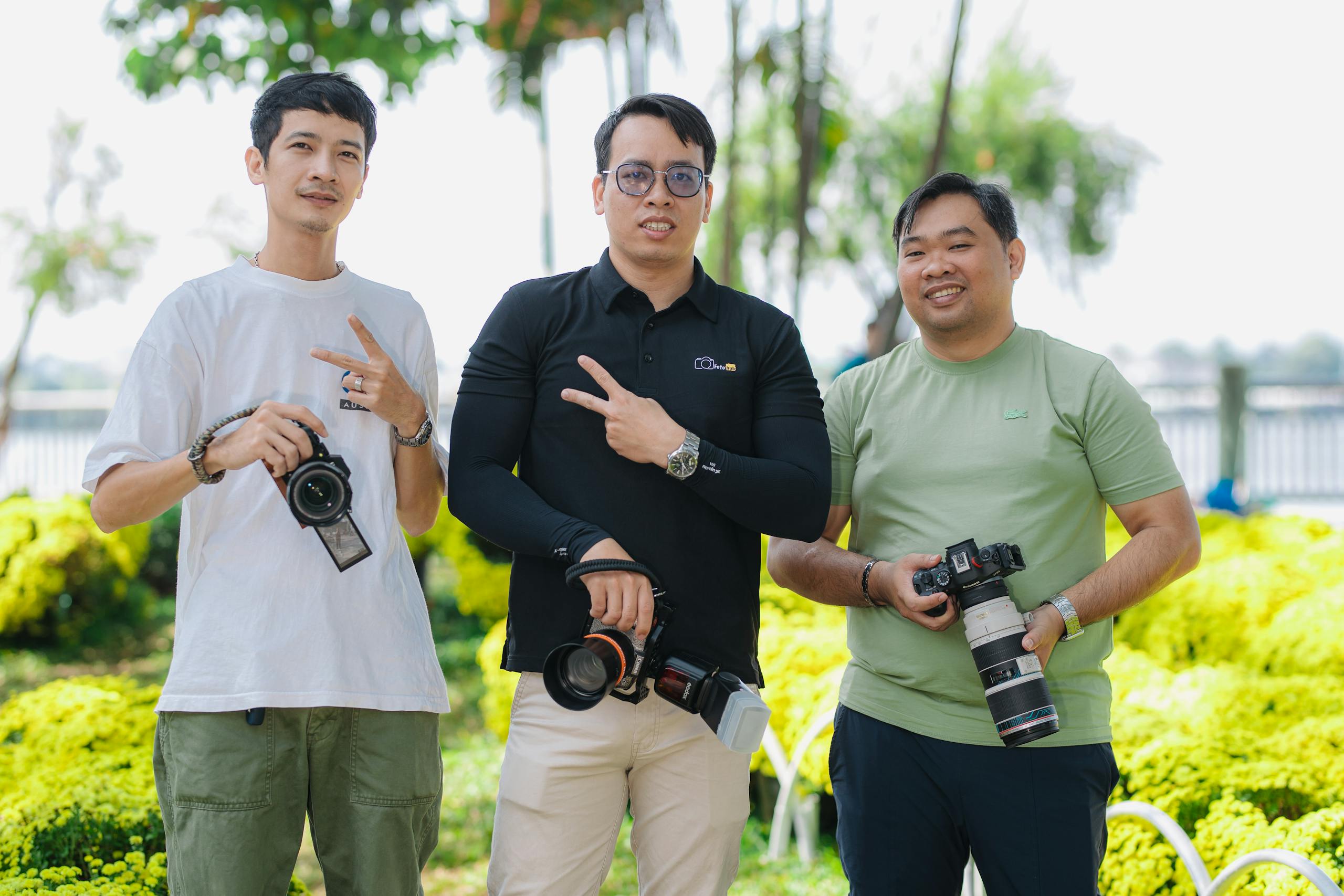 Three adult photographers displaying cameras in a sunny outdoor setting, showcasing relaxed friendship.